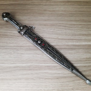 May include: A silver and black dagger with an ornate design and red gemstones. The dagger has a pointed tip and a handle with a decorative ring.