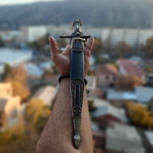 Pode incluir: Uma bainha de couro preto com detalhes prateados intrincados e uma ponta pontiaguda. A bainha &eacute; segurada em uma m&atilde;o com uma pulseira preta.