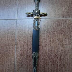 Pode incluir: Uma adaga decorativa com cabo preto e detalhes dourados. A l&acirc;mina &eacute; ornamentada e apresenta desenhos intrincados.