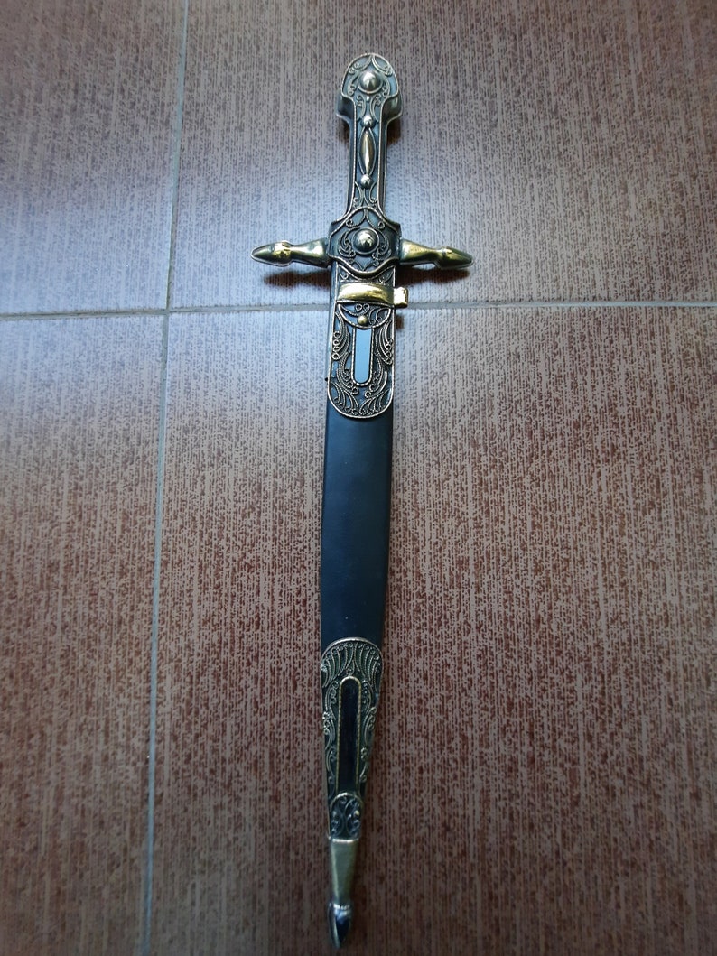 Pode incluir: Uma adaga decorativa com cabo preto e detalhes dourados. A l&acirc;mina &eacute; ornamentada e apresenta desenhos intrincados.