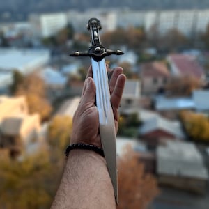 Pode incluir: Uma espada prateada com cabo preto e detalhes ornamentados. A espada &eacute; segurada em uma m&atilde;o com uma pulseira preta.