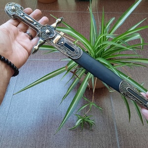 Pode incluir: Uma adaga decorativa de prata e ouro com uma bainha de couro preto. A adaga tem intrincados desenhos florais e uma ponta pontiaguda. A bainha tem uma ponta de metal decorativa.
