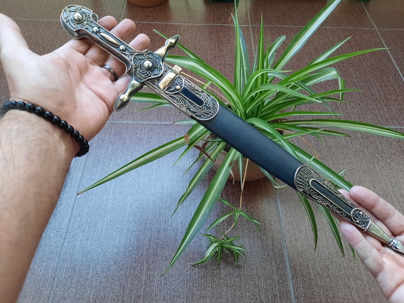 Pode incluir: Uma adaga decorativa de prata e ouro com uma bainha de couro preto. A adaga tem intrincados desenhos florais e uma ponta pontiaguda. A bainha tem uma ponta de metal decorativa.