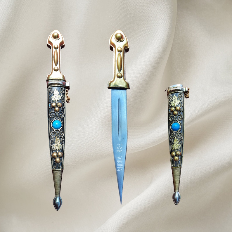 May include: Three decorative knives with gold and silver accents. The blades are silver and the handles are gold with intricate designs. The knives have blue gemstones on the handles. The middle knife has the text "FOR VIKING" engraved on the blade.