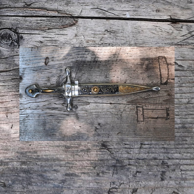 May include: A silver dagger with a black and gold handle and intricate details. The dagger is lying on a wooden surface.
