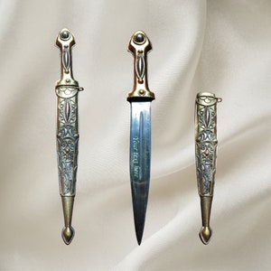 May include: Three ornate silver and gold daggers with intricate designs. The middle dagger has the text "Your text here" engraved on the blade.