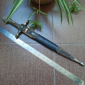 Pode incluir: Uma adaga decorativa com cabo preto e detalhes dourados. A adaga tem 48 cm de comprimento e possui um design detalhado no cabo.