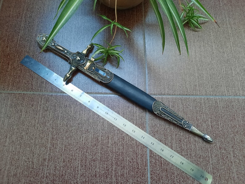 Pode incluir: Uma adaga decorativa com cabo preto e detalhes dourados. A adaga tem 48 cm de comprimento e possui um design detalhado no cabo.