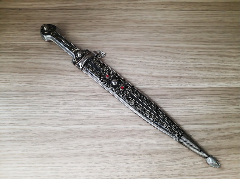 May include: A silver and black dagger with an ornate design and red gemstones. The dagger has a pointed tip and a handle with a decorative ring.
