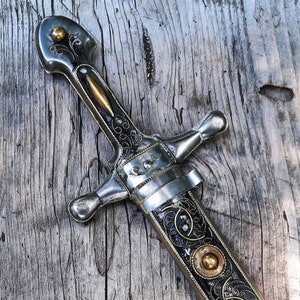 May include: A silver dagger with intricate floral designs and gold accents. The dagger has a pointed tip and a curved handle. The blade is decorated with a black and gold pattern.