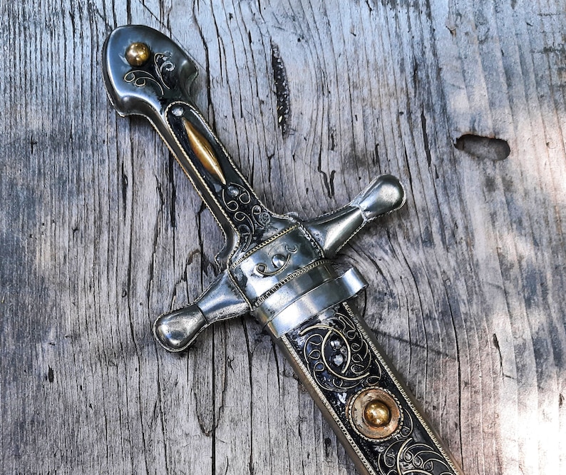 May include: A silver dagger with intricate floral designs and gold accents. The dagger has a pointed tip and a curved handle. The blade is decorated with a black and gold pattern.