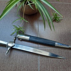 Pode incluir: Uma adaga de prata com uma bainha de couro preto. A adaga tem um cabo em forma de cruz e uma l&acirc;mina afiada. A bainha &eacute; decorada com uma faixa de prata.