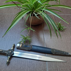 Pode incluir: Uma espada decorativa de prata e ouro com uma bainha de couro preto. A espada tem um cabo detalhado e uma l&acirc;mina afiada. A bainha tem entalhes intrincados e um acento dourado.