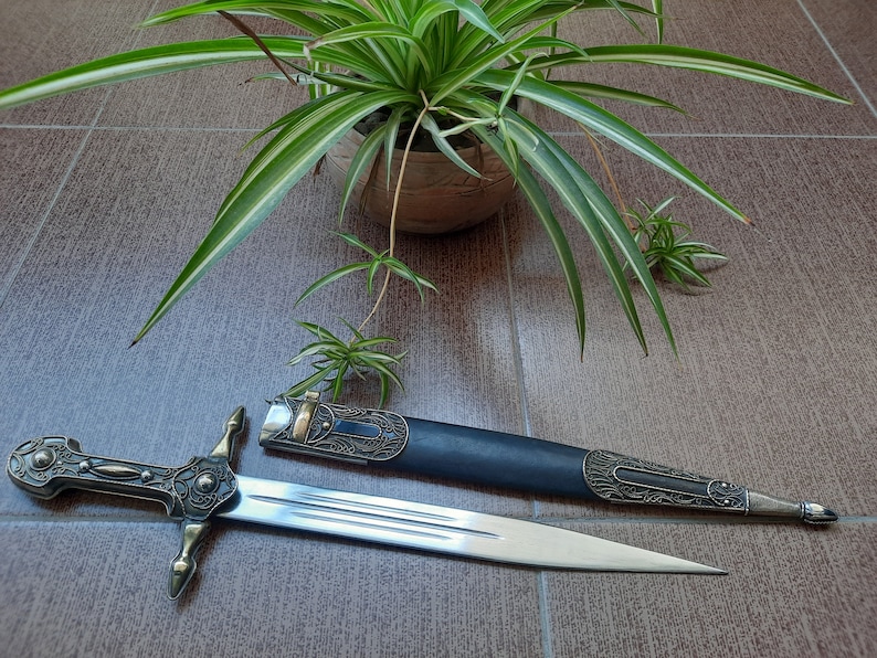 Pode incluir: Uma espada decorativa de prata e ouro com uma bainha de couro preto. A espada tem um cabo detalhado e uma l&acirc;mina afiada. A bainha tem entalhes intrincados e um acento dourado.