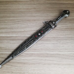 May include: A silver dagger with an intricate black and gold design. The dagger has a red jewel on the handle.