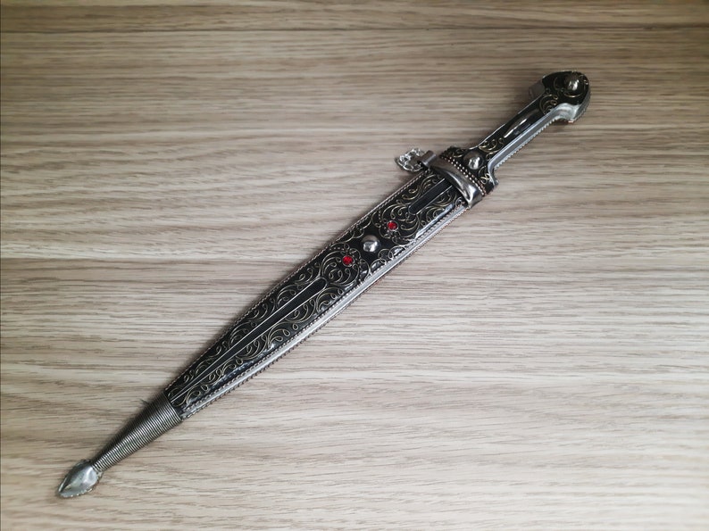 May include: A silver dagger with an intricate black and gold design. The dagger has a red jewel on the handle.
