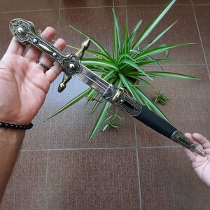 Pode incluir: Uma adaga prateada e preta com detalhes ornamentados e um cabo preto. A adaga est&aacute; sendo segurada em uma m&atilde;o e est&aacute; apoiada em uma planta verde.