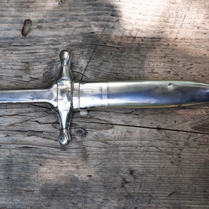 May include: A silver dagger with a cross-shaped handle and a pointed blade. The dagger is lying on a wooden surface.