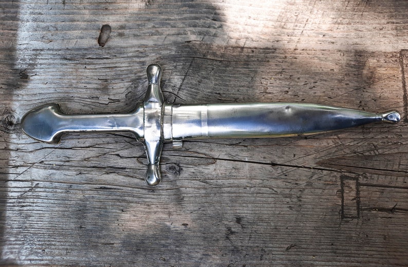 May include: A silver dagger with a cross-shaped handle and a pointed blade. The dagger is lying on a wooden surface.