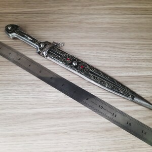 May include: A silver and black decorative dagger with intricate floral designs and red gemstones. The dagger is 14 inches long and is measured against a ruler.