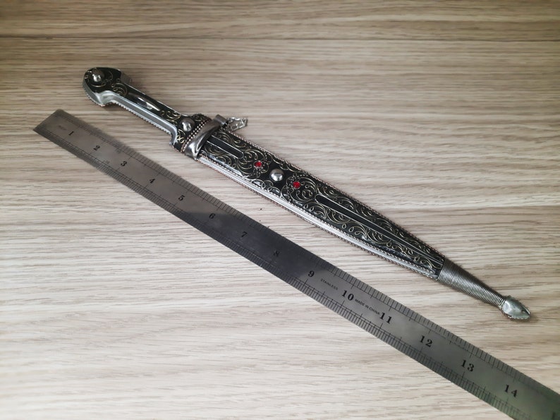 May include: A silver and black decorative dagger with intricate floral designs and red gemstones. The dagger is 14 inches long and is measured against a ruler.
