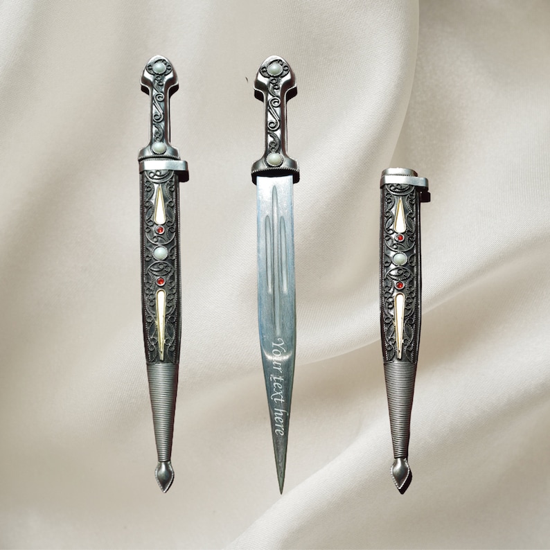 May include: Three silver decorative daggers with intricate designs and red gemstones. The daggers have a pointed tip and a curved handle.