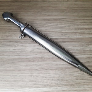 May include: A silver dagger with a decorative handle and a pointed tip. The dagger is lying on a light brown wooden surface.