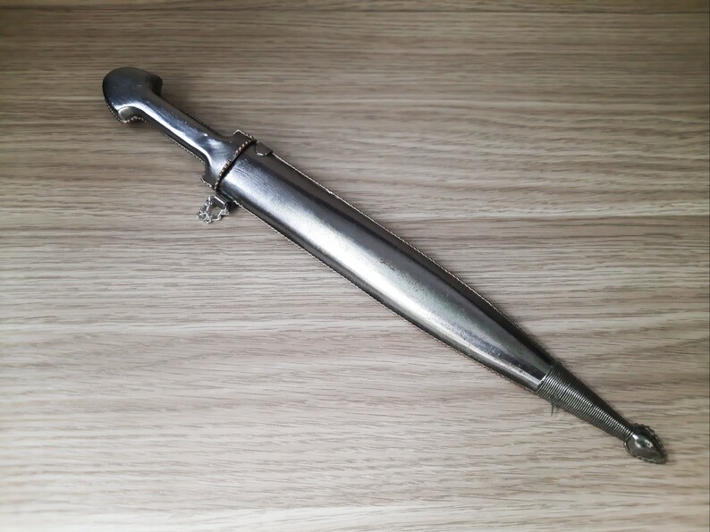 May include: A silver dagger with a decorative handle and a pointed tip. The dagger is lying on a light brown wooden surface.