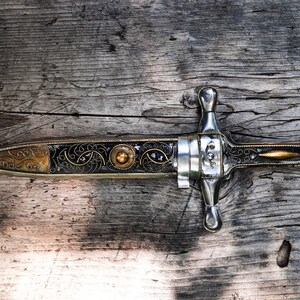 May include: A silver and gold dagger with intricate floral designs on a wooden surface.