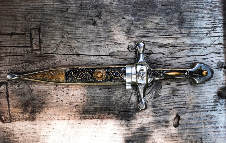 May include: A silver and gold dagger with intricate floral designs on a wooden surface.