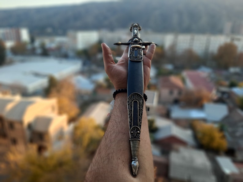 Pode incluir: Uma bainha de couro preto com detalhes prateados intrincados e uma ponta pontiaguda. A bainha &eacute; segurada em uma m&atilde;o com uma pulseira preta.