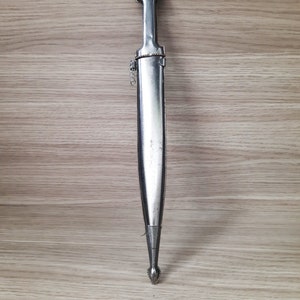 May include: A silver-colored dagger with a pointed tip and a decorative handle. The dagger is displayed against a woodgrain background.