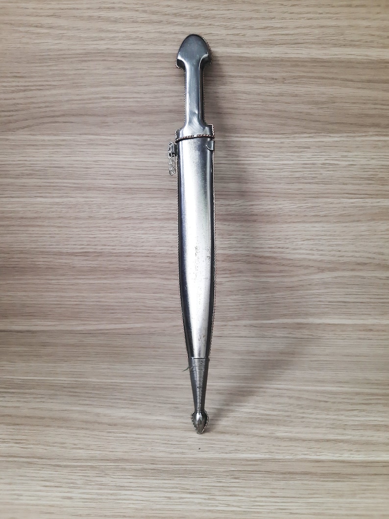May include: A silver-colored dagger with a pointed tip and a decorative handle. The dagger is displayed against a woodgrain background.