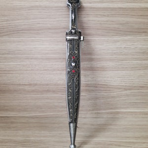 May include: A silver and black dagger with intricate floral designs and red gemstones. The dagger has a pointed tip and a decorative handle.