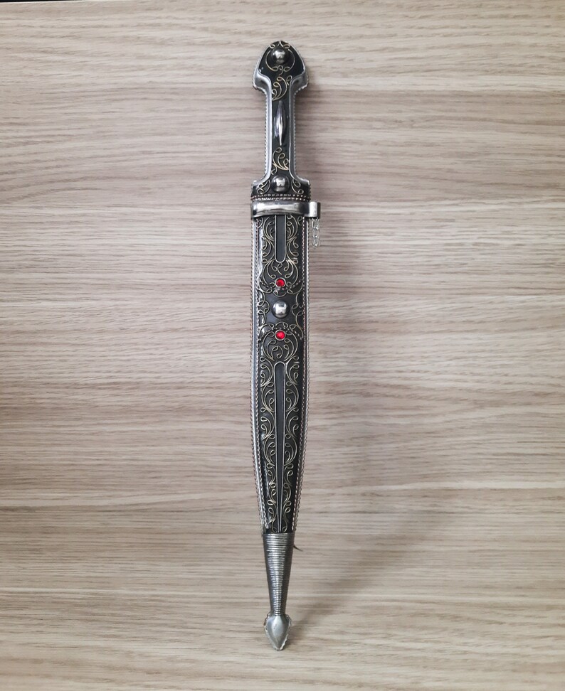 May include: A silver and black dagger with intricate floral designs and red gemstones. The dagger has a pointed tip and a decorative handle.