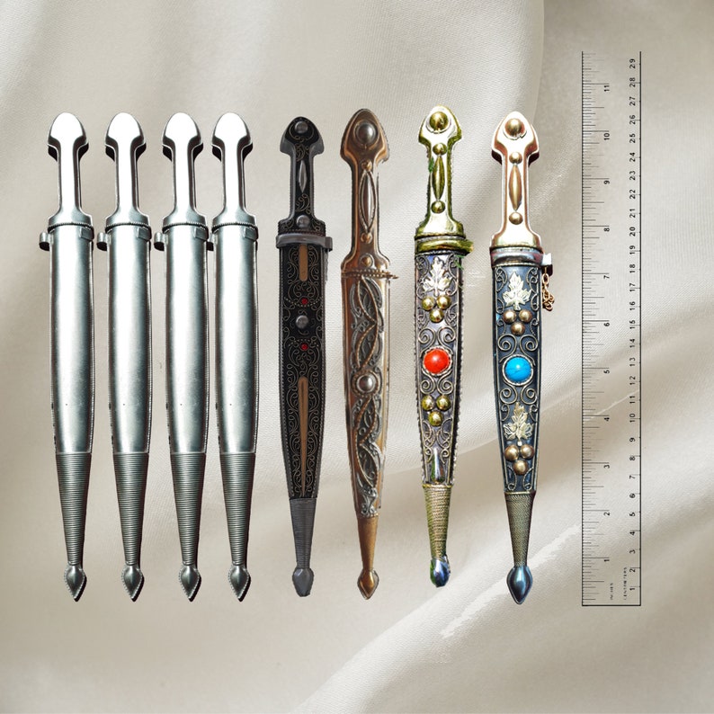 May include: Seven decorative daggers with intricate designs. The daggers have silver, gold, and black finishes with colorful accents. The daggers are arranged in a row on a white fabric background.