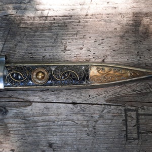 May include: A silver and gold dagger with intricate floral designs on the handle. The dagger is lying on a wooden surface.