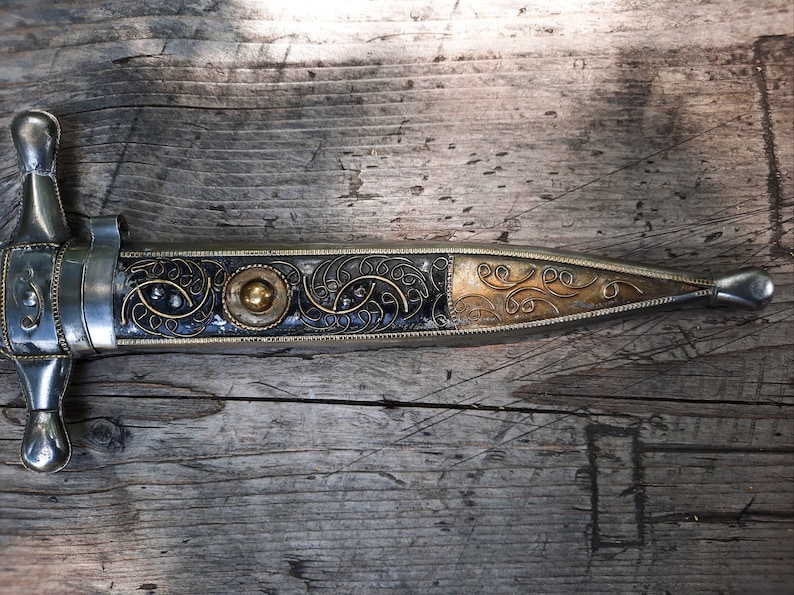 May include: A silver and gold dagger with intricate floral designs on the handle. The dagger is lying on a wooden surface.