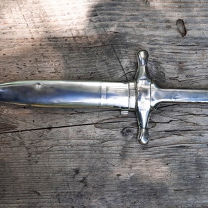May include: A silver dagger with a pointed tip and a decorative handle with two round knobs. The dagger is lying on a wooden surface.