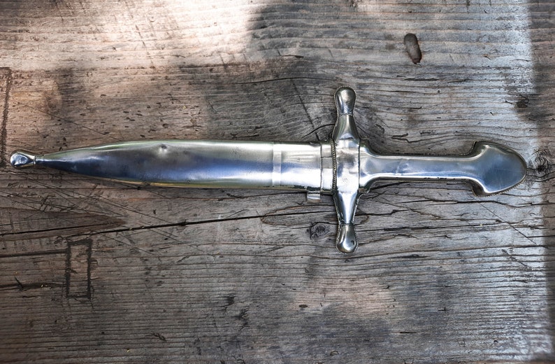 May include: A silver dagger with a pointed tip and a decorative handle with two round knobs. The dagger is lying on a wooden surface.