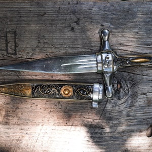 May include: A silver dagger with a decorative handle and a matching sheath. The dagger has a gold-coloured accent on the handle and a gold-coloured band on the sheath. The dagger and sheath are lying on a wooden surface.