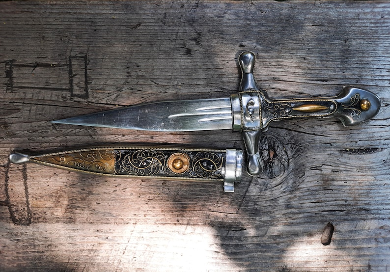 May include: A silver dagger with a decorative handle and a matching sheath. The dagger has a gold-coloured accent on the handle and a gold-coloured band on the sheath. The dagger and sheath are lying on a wooden surface.