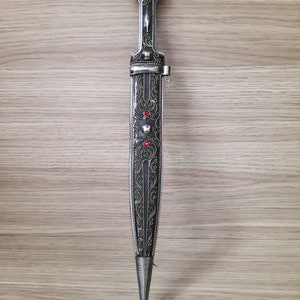 May include: A silver dagger with intricate floral engravings and red gemstones. The dagger has a black handle and a pointed tip.
