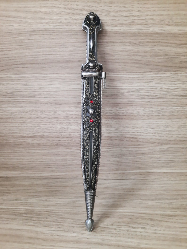 May include: A silver dagger with intricate floral engravings and red gemstones. The dagger has a black handle and a pointed tip.