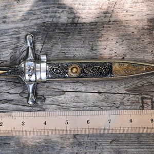 May include: A silver and gold dagger with an ornate black handle and a blade that is approximately 23 cm long. The dagger is resting on a wooden surface.