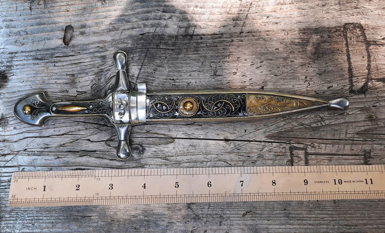 May include: A silver and gold dagger with an ornate black handle and a blade that is approximately 23 cm long. The dagger is resting on a wooden surface.