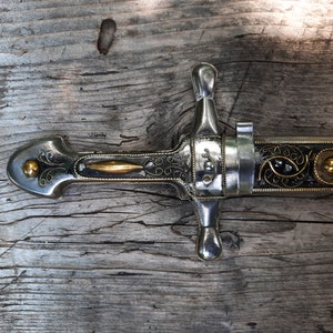 May include: A silver and gold decorated dagger with a black handle and intricate details. The dagger is lying on a wooden surface.