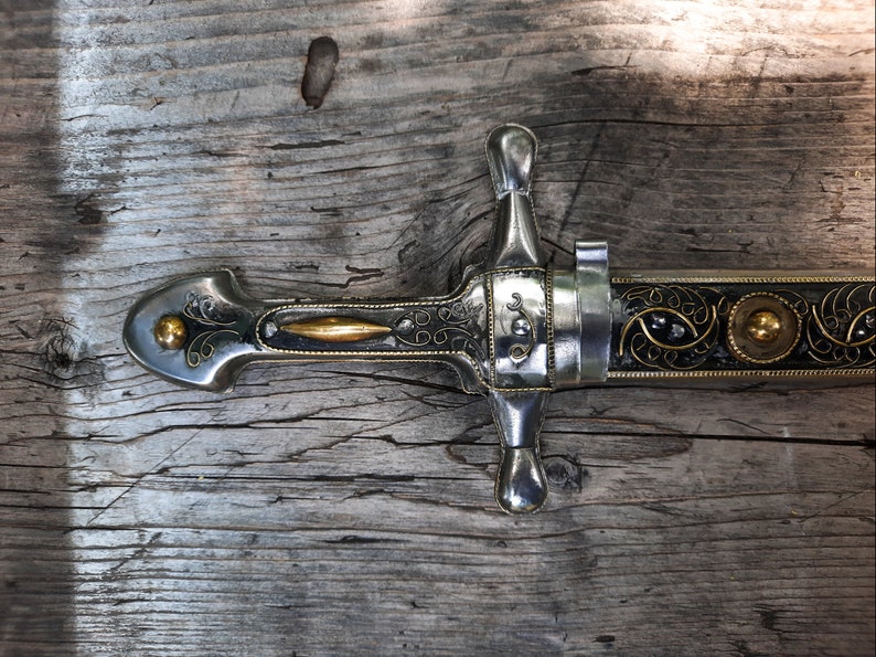 May include: A silver and gold decorated dagger with a black handle and intricate details. The dagger is lying on a wooden surface.