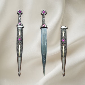May include: Three silver daggers with ornate designs and pink gemstones. The middle dagger has the inscription "To Iris My Friend".