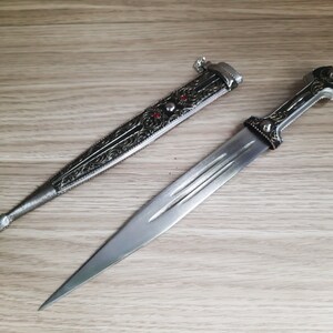 May include: A silver dagger with an ornate handle and a black sheath. The dagger has a sharp point and a double-edged blade.